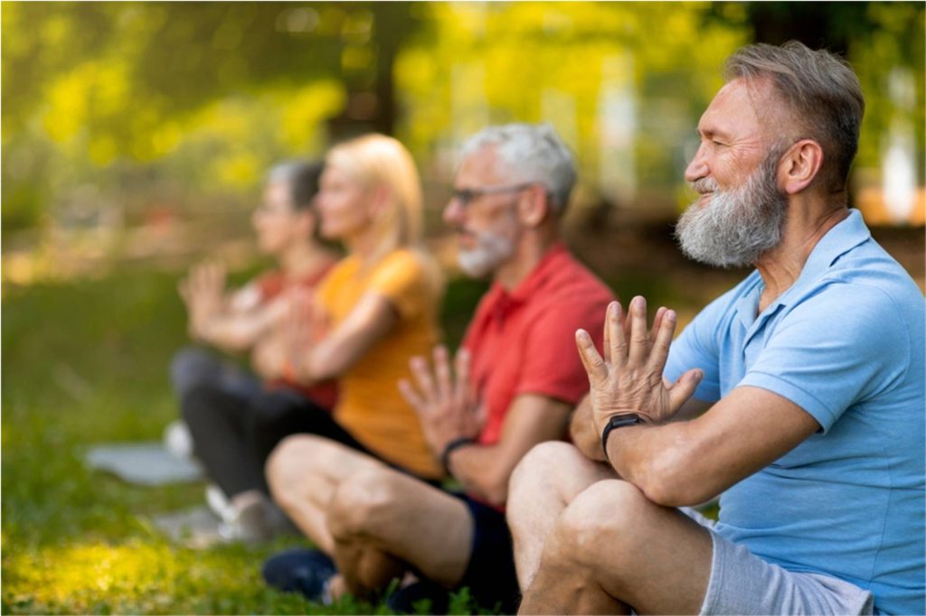 Group of older adults sitting cross-legged outdoors, practicing yoga with hands in a prayer position, enjoying a mindful and active lifestyle.