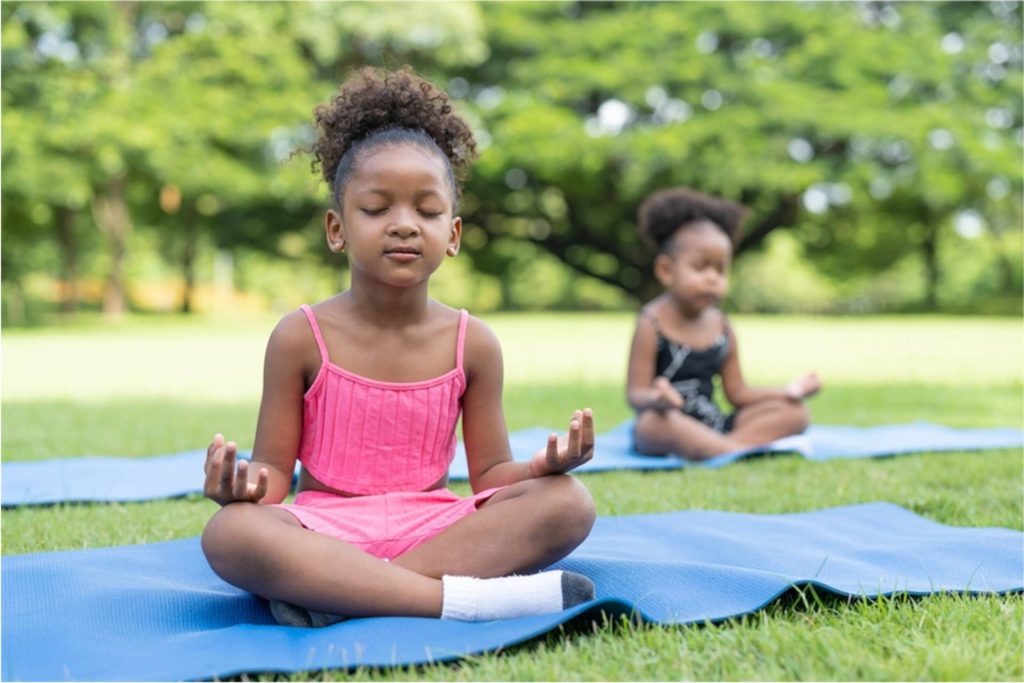 Young girl sitting cross-legged on a blue yoga mat outdoors, eyes closed, practicing meditation in a kids’ yoga class.